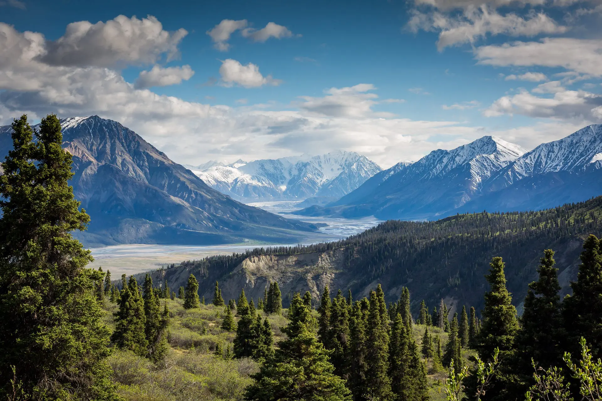 A wide mountain valley scene with snow-capped peaks and a distant glacier under a partly cloudy blue sky, evergreen trees and green foothills in the foreground suggesting stability, safety, and a long-term, protected retirement outlook