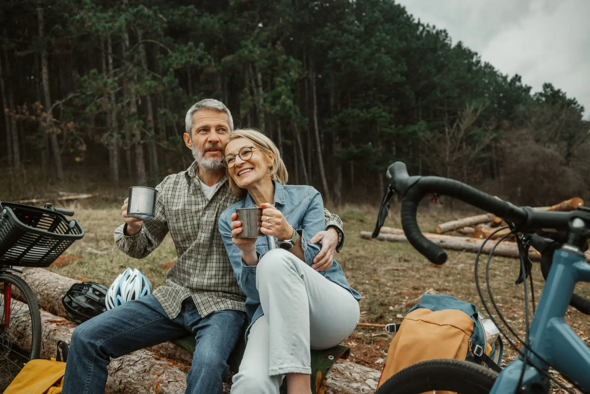 Mature cycling couple enjoying coffee break in nature Older couple sitting on a log in a forest clearing, holding metal mugs and leaning together with bicycles, helmets, and a backpack nearby, conveying relaxed retirement, outdoor freedom, and safety-first planning.